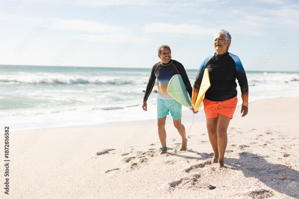 Obraz premium Full length of happy multiracial senior couple walking with surfboards on sand at sunny beach