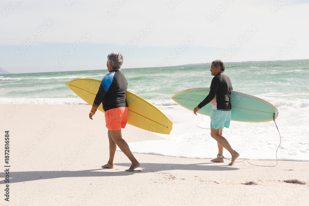 Obraz premium Full length of multiracial senior couple carrying surfboard walking on shore at sunny beach