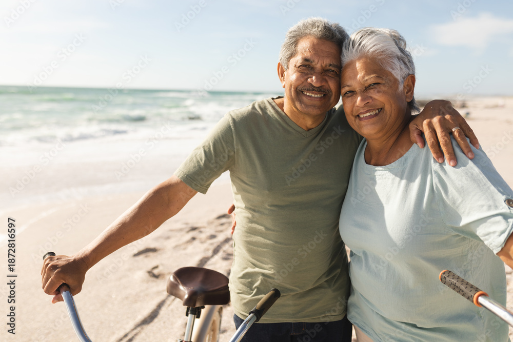 Fototapeta premium Portrait of smiling senior multiracial couple with arm around bicycles at beach on sunny day