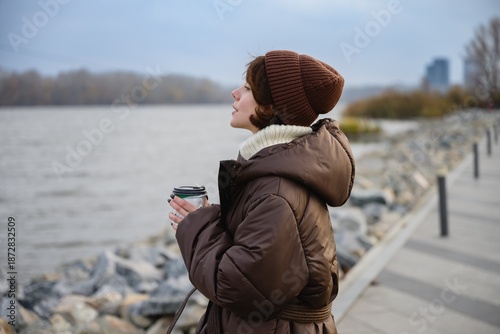 Thoughtful woman in a warm jacket and knit hat holding a takeaway coffee while standing by a river embankment. Calm autumn mood, slow living, urban nature and peaceful outdoor lifestyle.