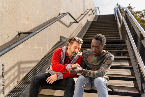 Two friends of different ethnicities sitting on the stairs in the city, looking at a bracelet and talking