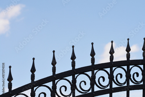 The upper part of a black wrought iron fence featuring ornate swirls and pointed spikes stands against a light blue sky with soft white clouds