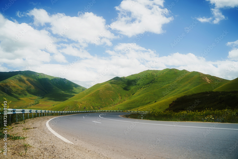 Obraz premium Winding asphalt road through lush green mountains under a bright blue sky with fluffy white clouds