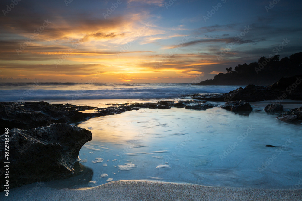 Obraz premium A golden sun setting over a rocky shoreline on an Indonesian island. Long exposure capture on sunset featuring a shallow blue tidal pool below vibrant orange clouds.