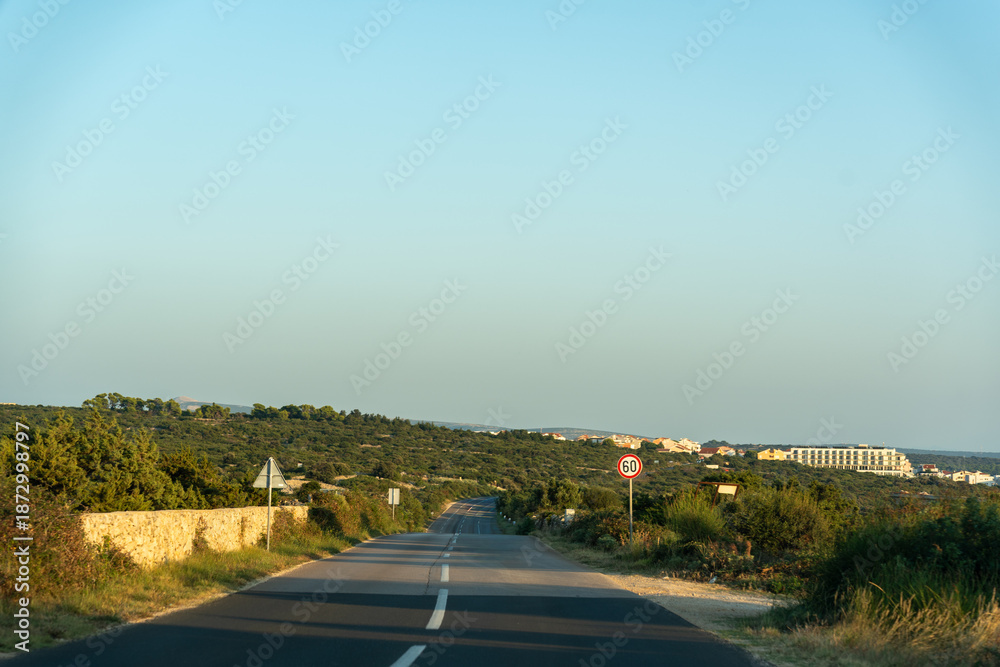 Fototapeta premium Empty country road with 60 km speed limit sign leading to coastal town through green Mediterranean hills on summer evening. Concept of road trip travel, scenic route, driving journey, vacation drive.