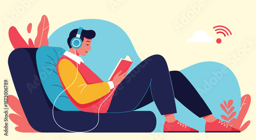 Young man relaxes on a blue sofa while immersed in a book and listening to music through headphones in a cozy indoor setting.