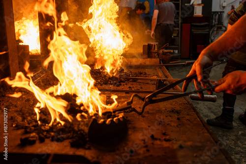Photos Scène de forge en atelier avec un artisan manipulant du métal au feu à l’aide de pinces, illustrant la puissance du travail traditionnel de ferronnerie