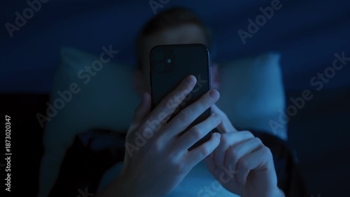 Close up of man with a smartphone in his hands, illuminated with blue light of the screen, browsing social media and consuming content late at night instead of sleeping. Digital addiction and insomnia