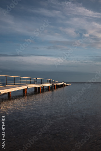 A quiet wooden pier stretching into calm sea water under a dramatic cloudy sky, creating a peaceful and minimal coastal scene.