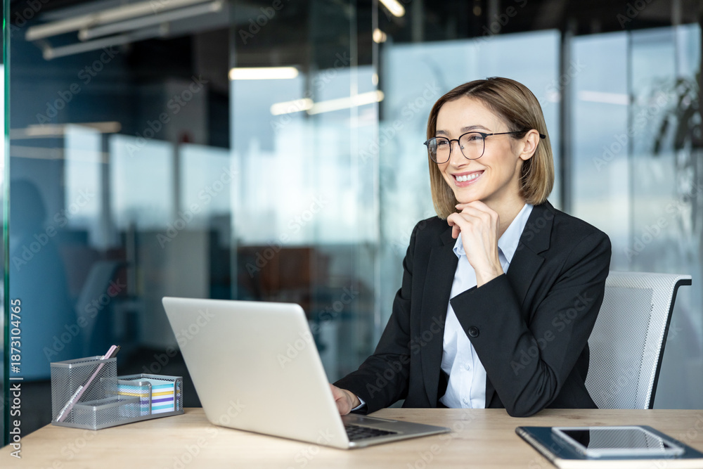 Fototapeta premium Young businesswoman wearing eyeglasses looking away and smiling while sitting at her modern office desk with a laptop. Embodying innovation. Success. And professional growth in a corporate environment