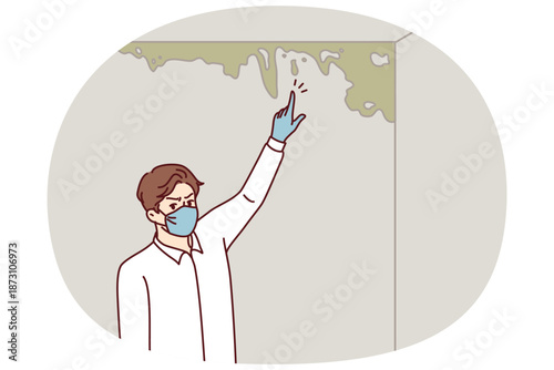 Man near wall with mold points finger at spreading fungus that has arisen due to violation of building codes. Inspector in white shirt and protective mask stands in room with mold.