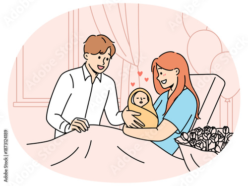 Mother with newborn baby sits on couch in hospital and shows infant to happy father. Smiling married couple have become parents for first time admire sleeping newborn wrapped in blanket