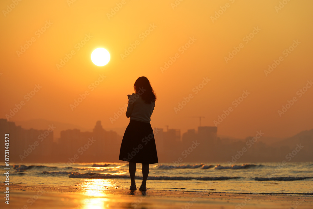 Fototapeta premium Silhouette of woman standing with smartphone on sea beach on city background. Sunrise on a coast, travel and holiday concept