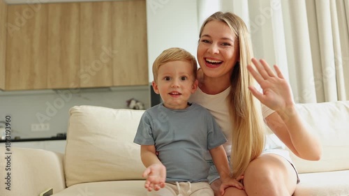 Blonde mother and her adorable son wave cheerfully, then embrace with smiles and a sweet kiss in the living room