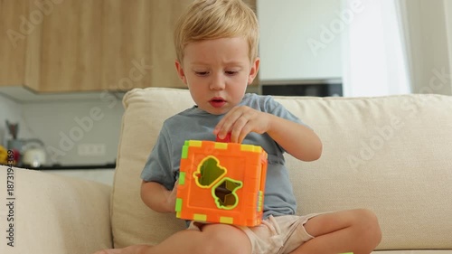 A curious toddler boy sits comfortably on a sofa, play with in a vibrant shape sorter toy. developing problem-solving skills, carefully placing blocks into matching holes