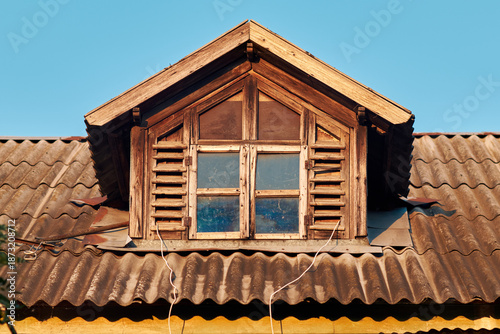 Rustic wooden attic window with shutters on old rooftop under clear blue sky