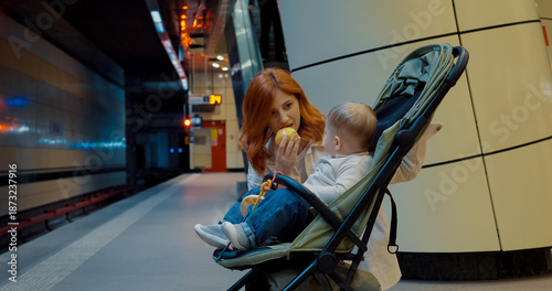 Принти на полотні Young mother feeds her baby an apple while waiting for the subway train