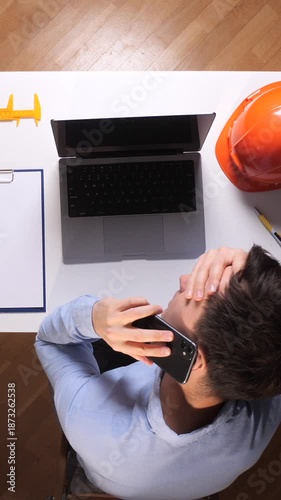 Vertical video engineer man covering his face with his hand feeling stressed and frustrated during a phone call, working with a laptop and safety helmet on a white desk