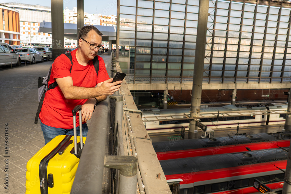 Fototapeta premium Solo male traveler waiting at a modern railway station, holding a yellow suitcase and checking his phone while looking over the train platforms below.
