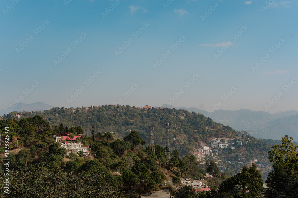 Fototapeta premium Scenic Mountain Landscape with Lush Greenery and a Clear Blue Sky, Serene Vista of Rolling Hills and Scattered Buildings, Perfect for Travel and Nature