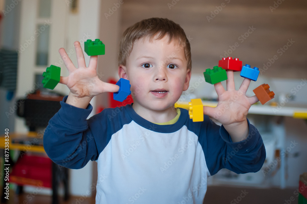 Fototapeta premium A young boy holds various small building blocks on his fingers in a room filled with toys. He smiles while playing and enjoying his time with colorful toys