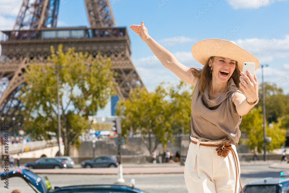 Fototapeta premium A confident woman in a wide-brimmed straw hat captures a vibrant selfie in front of the iconic Eiffel Tower on a sunny day, celebrating her solo travel adventure in Paris.