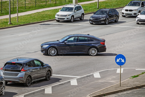 Urban traffic scene viewed from an elevated angle, showing several passenger cars driving through a multi-lane intersection with road markings and traffic signs on a clear day