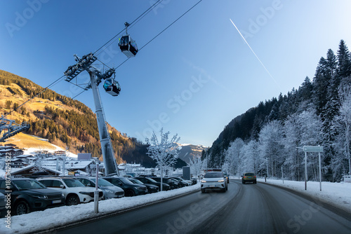 Εκτύπωση καμβά Scenic alpine village resort in Austria gondola cablecar above snowy road car parking lots trees mountain valley scenery under clear blue winter sky