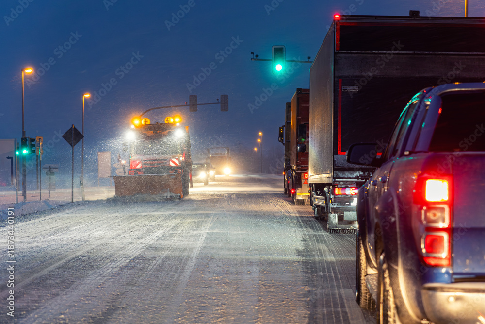 Fototapeta premium Night winter road scene heavy snowfall snowplow truck clearing icy street, cars freight vehicles waiting green traffic light extreme cold weather conditions. Dangerous urban transport at blizzard