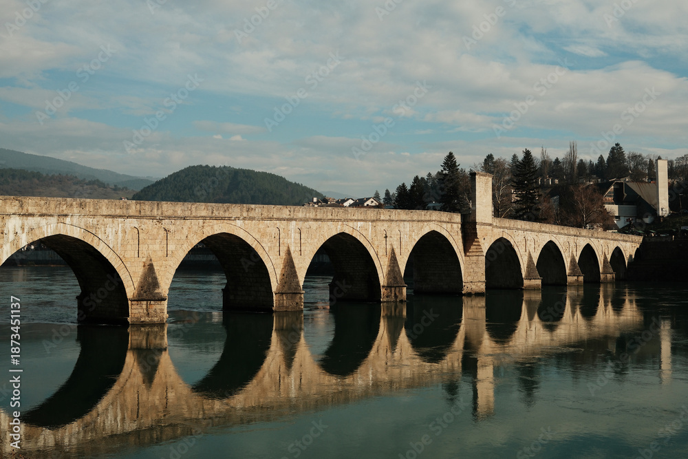 Fototapeta premium Historic Mehmed Pasha Sokolovic Bridge spans the Drina River in Visegrad, Bosnia and Herzegovina. Stone arches and soft reflections highlight the calm atmosphere of this iconic landmark.