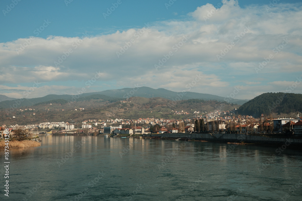 Fototapeta premium Wide view of the Drina River flowing past Visegrad with hills and residential buildings along the riverbank. Calm water and open sky emphasize the natural rhythm of the town.