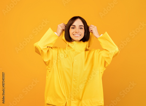 Photos Happy woman in yellow waterproof raincoat putting on hood, standing isolated on yellow background and smiling to camera