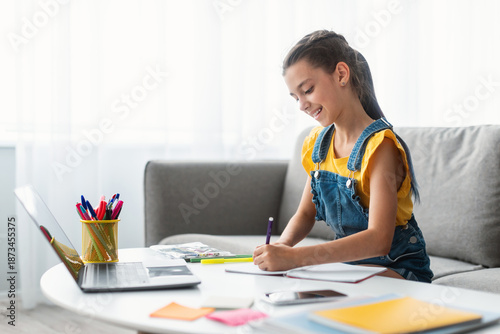 Tableau sur toile Portrait of smiling schoolgirl sitting on couch at desk with pc, writing essay or drawing picture in paper notebook