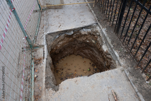 Deep hole excavated in a concrete sidewalk, filled with muddy water and fallen leaves, next to a temporary construction fence