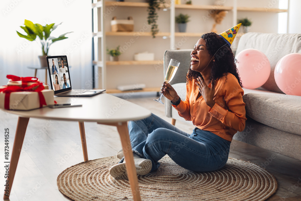 Obraz premium Distance Birthday Party. Joyful Black Lady At Laptop Celebrating B-Day Online Drinking Champagne Having Group Video Call With Friends Sitting At Home, Wearing Festive Hat. Side View Shot