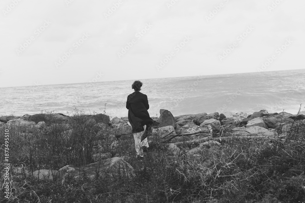 Obraz premium Solitude on a rocky shoreline as a lone figure walks toward the sea, creating a moody seascape with textured rocks, calm water, and a distant horizon in grayscale
