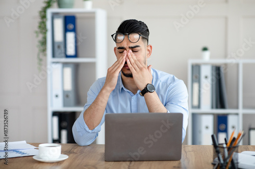 Fotografía Millennial Arab employee rubbing tired irritated eyes, working with laptop too much, exhausted from online job at office