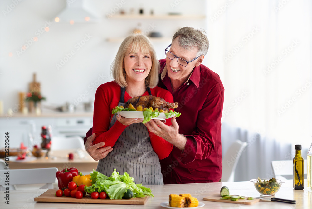 Fototapeta premium Positive Caucasian senior couple cooking Thanksgiving or Christmas turkey together, preparing traditional meal for family celebration at kitchen. Festive occasion concept