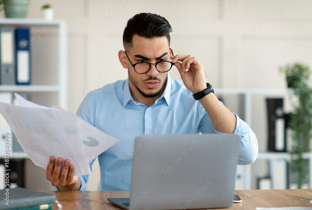 Obraz premium Stressed young Arab employee using laptop, holding documents, having problem with work task, making mistake in business project at office. Millennial guy having trouble meeting deadline