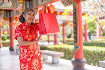 Senior asian woman in red cheongsam happily holding festive shopping bags at Chinese temple during Lunar New Year, festive shopping, tradition, celebration and cultural, CNY Campaign or flash sale