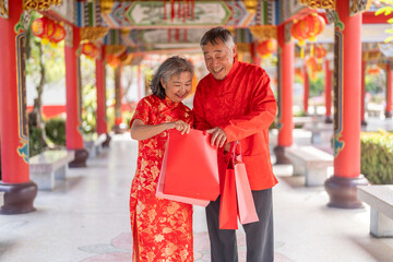 Happy senior Asian couple holding red shopping bags at Chinese temple during Lunar New Year, Elderly husband and wife laughing in cheongsam celebrating shopping festival. Retirement lifestyle concept
