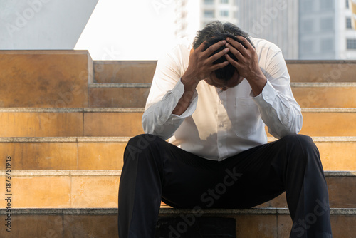 tressed Asian businessman sitting on city stairs holding head with hands. Depressed male office worker suffering from unemployment, bankruptcy or burnout failure concept.