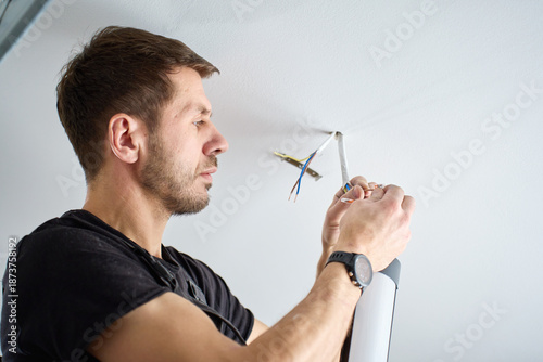 Billede på lærred Man in workwear connecting electrical wires while installing ceiling light fixture indoors