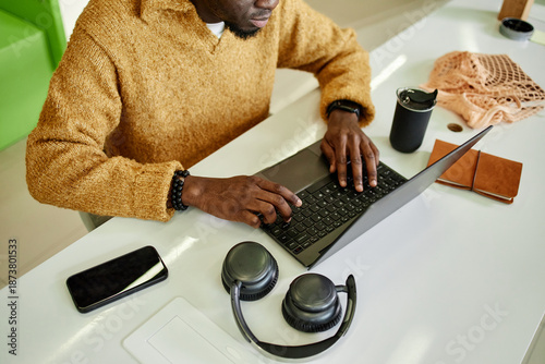 Young adult Black man working on laptop at desk, hands typing on keyboard, smartphone and wireless headphones nearby, reusable cup and notebook on workspace, partial face visible