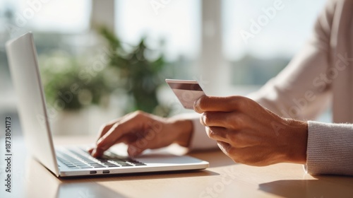 Hands are holding a credit card while typing on a laptop keyboard. The setting features natural daylight in a clean workspace with no distractions