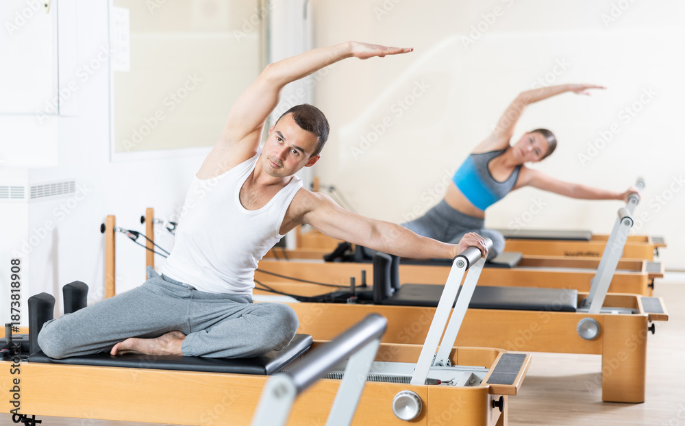 Fototapeta premium Concentrated athletic young man performing set of pilates exercises on reformer during group workout in fitness studio. Healthy active lifestyle..