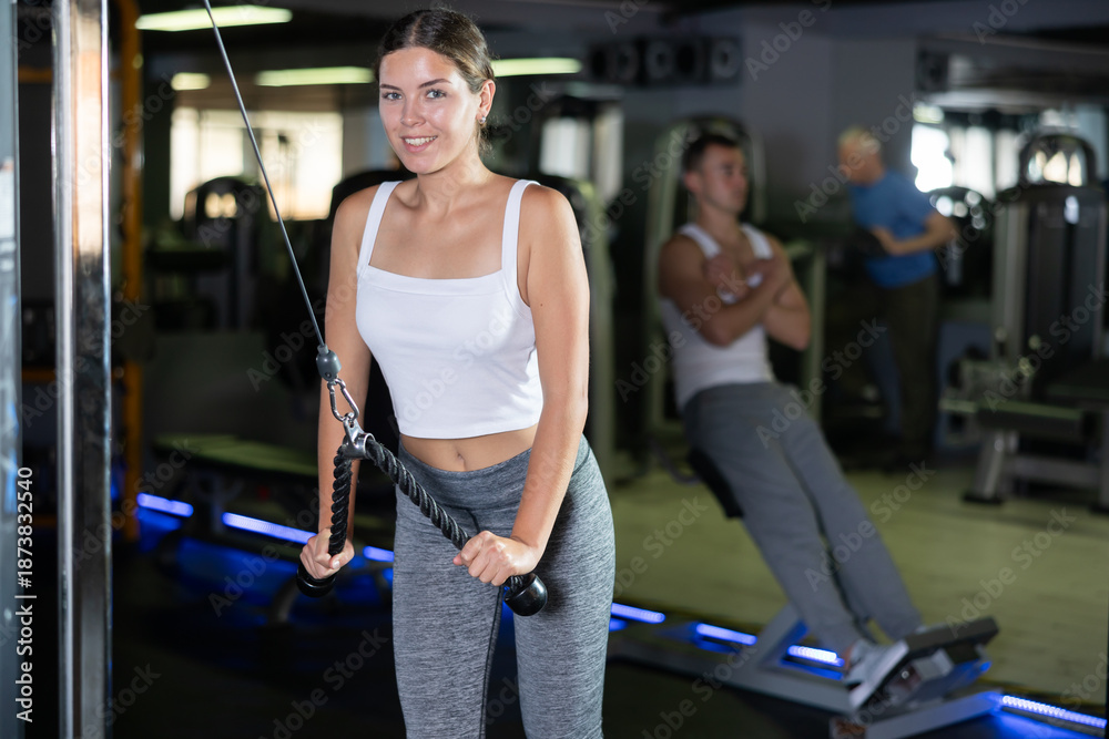 Fototapeta premium Slender athletic girl work out muscles of arms and shoulders on rope loop simulator in gym of fitness club