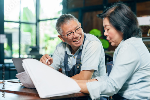 Elderly asian couple wearing apron smiling while reviewing document together at wooden table in restaurant discussing small business planning teamwork cooperation partnership expressing positive work