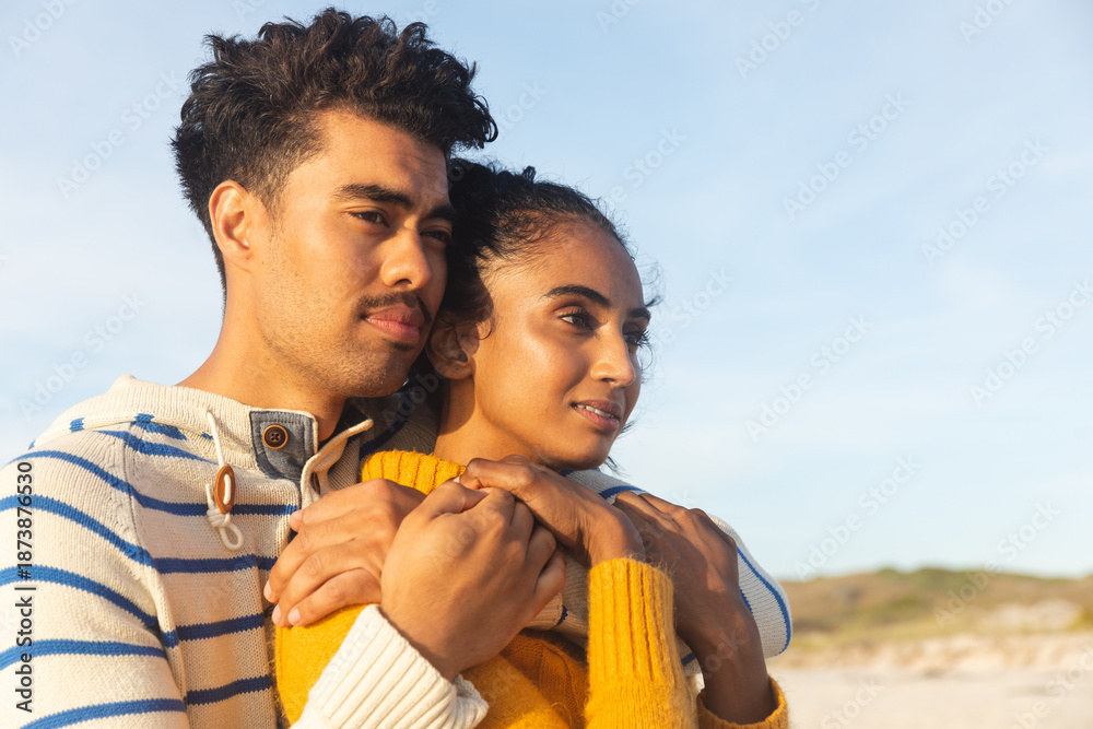 Fototapeta premium Thoughtful multiracial couple embracing while looking away at beach against sky on sunny day