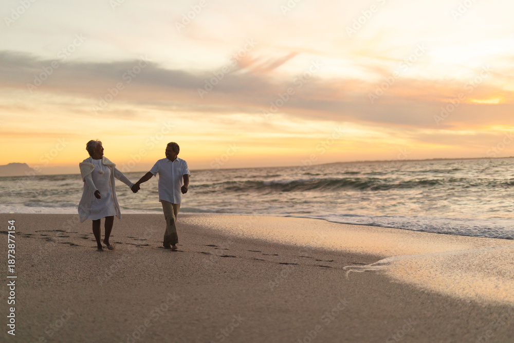 Fototapeta premium Senior couple holds hands on beach walk at sunset